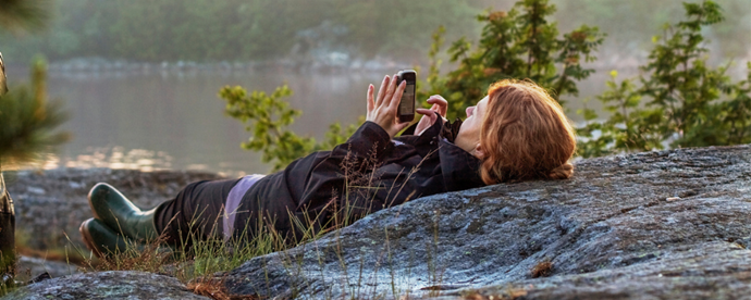 Person ligger på en klippa och tittar i en telefon. Foto.