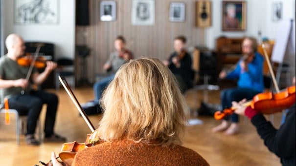 Kvinna som spelar fiol i musiksal i förgrunden. I bakgrunden fler som spela fiol.